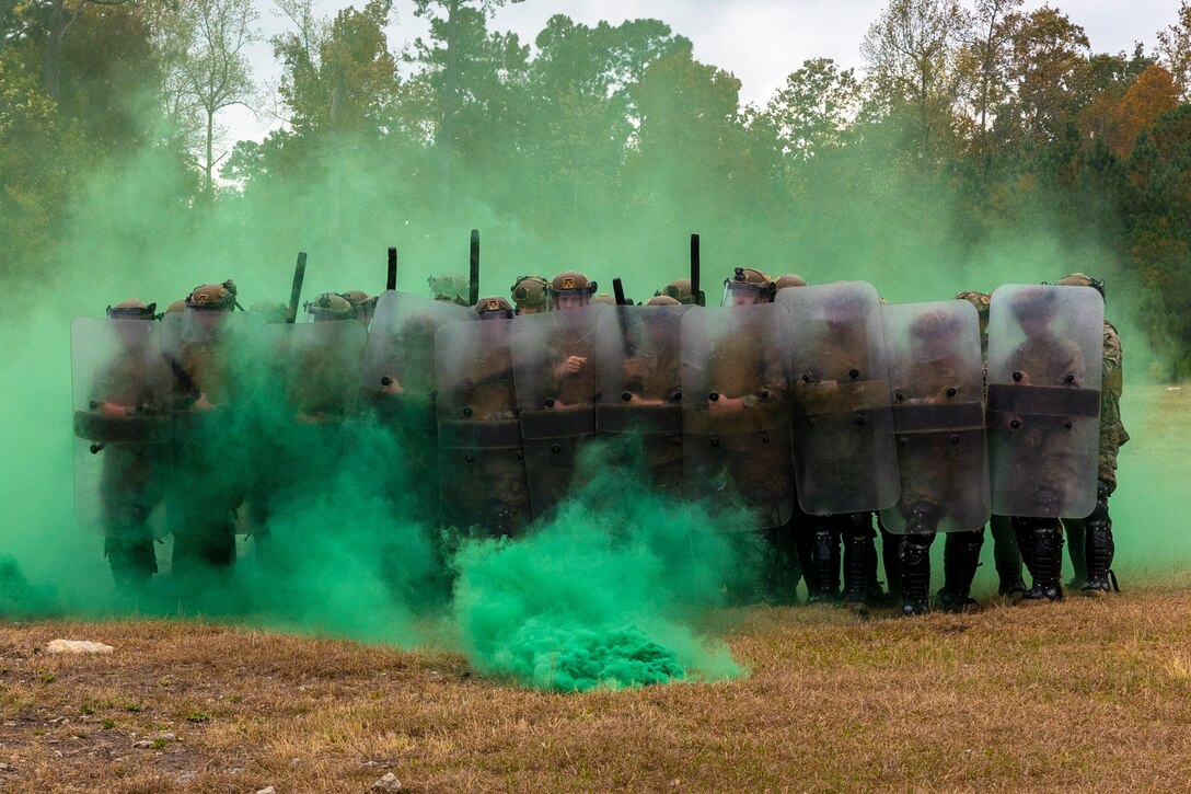 Marines holding clear shields stand in formation in a field surrounded by clouds of green smoke.