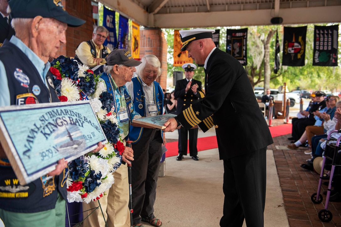 A sailor hands a gift o a veteran during an outdoor ceremony as others standing next to him look on.