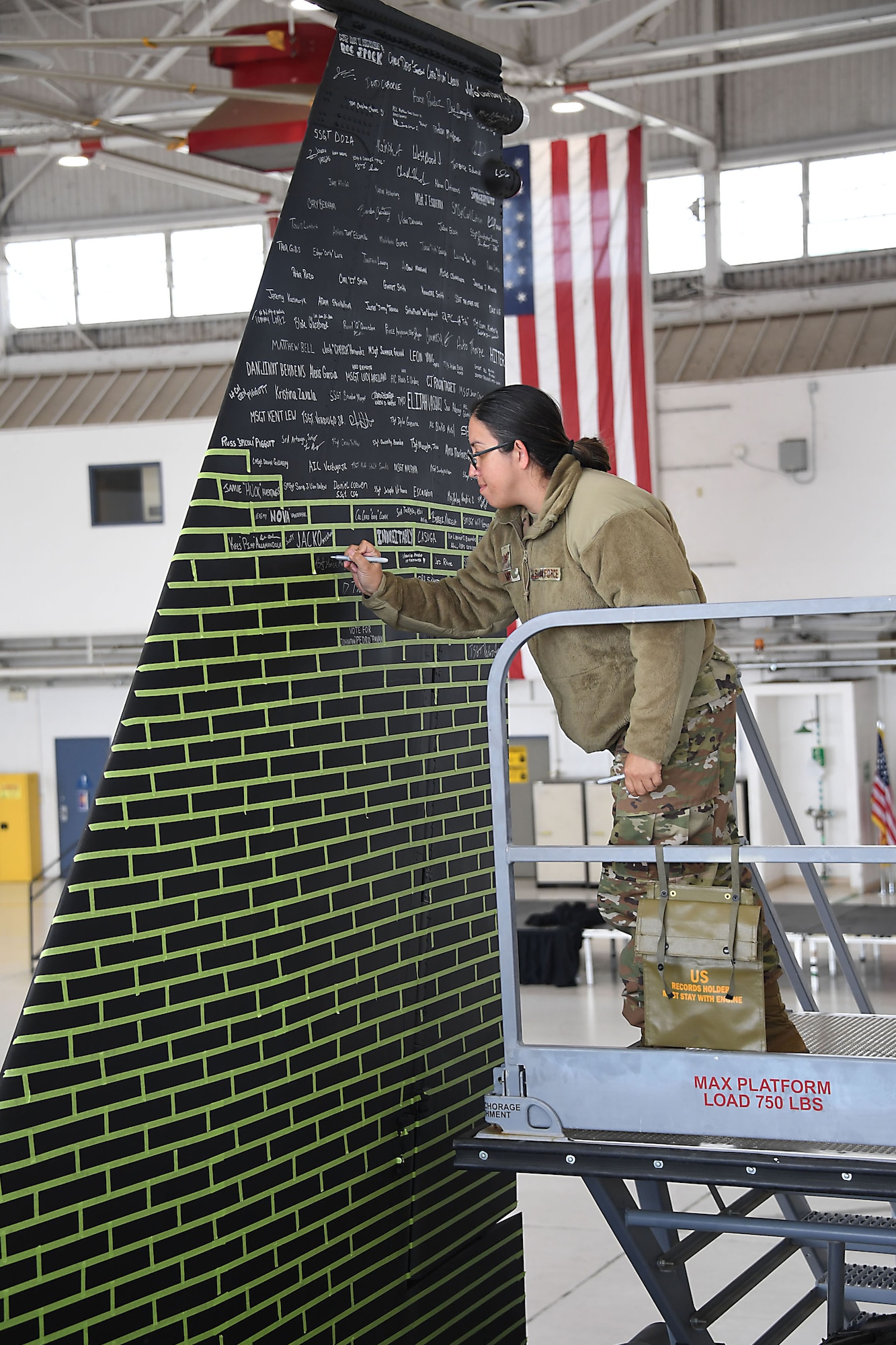 A woman wearing a military uniform stands on a ladder to sign the tail of an aircraft.