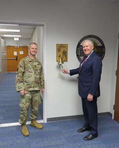Secretary of the Air Force rings a large wall mounted bell while standing next to officer in camouflaged uniform.