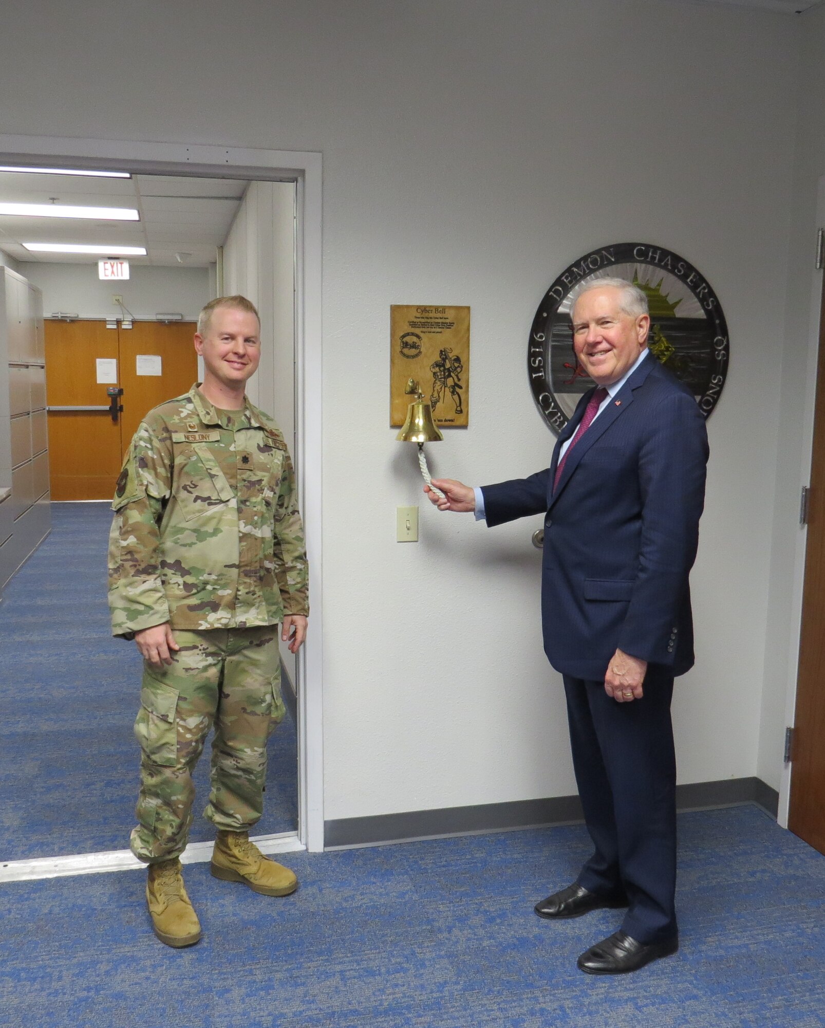Secretary of the Air Force rings a large wall mounted bell while standing next to officer in camouflaged uniform.