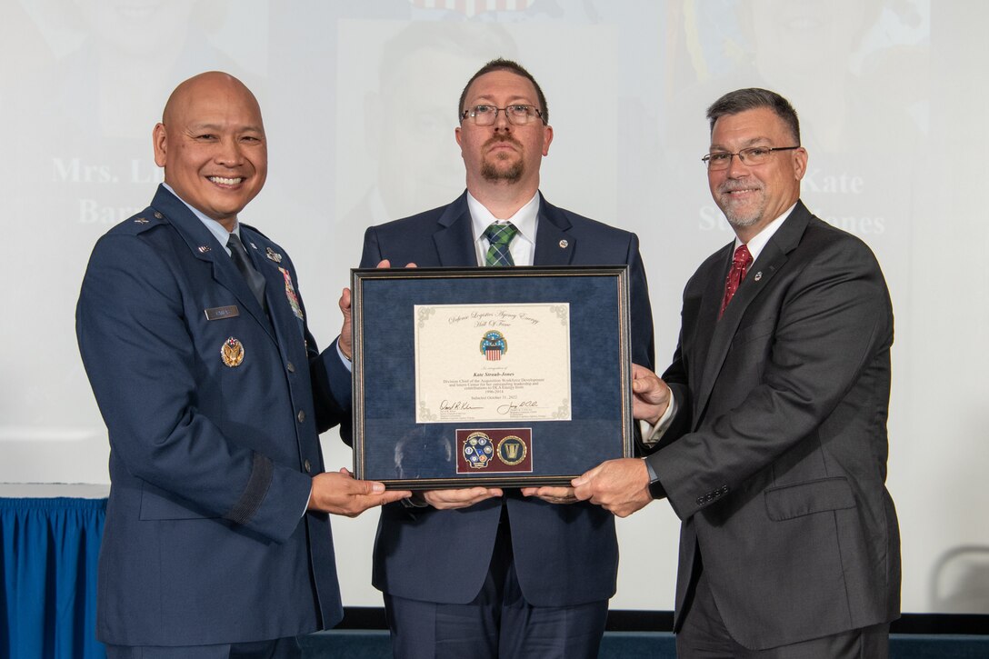 three gentlemen stand on a stage holding a plaque