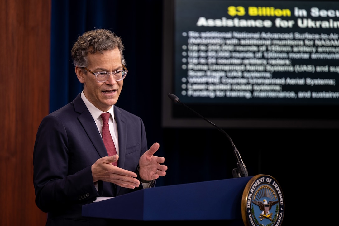 A man, standing at a lectern, gestures to the audience.