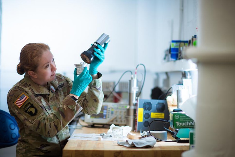 A woman in uniform wearing rubber gloves holds and observes an object in a laboratory.