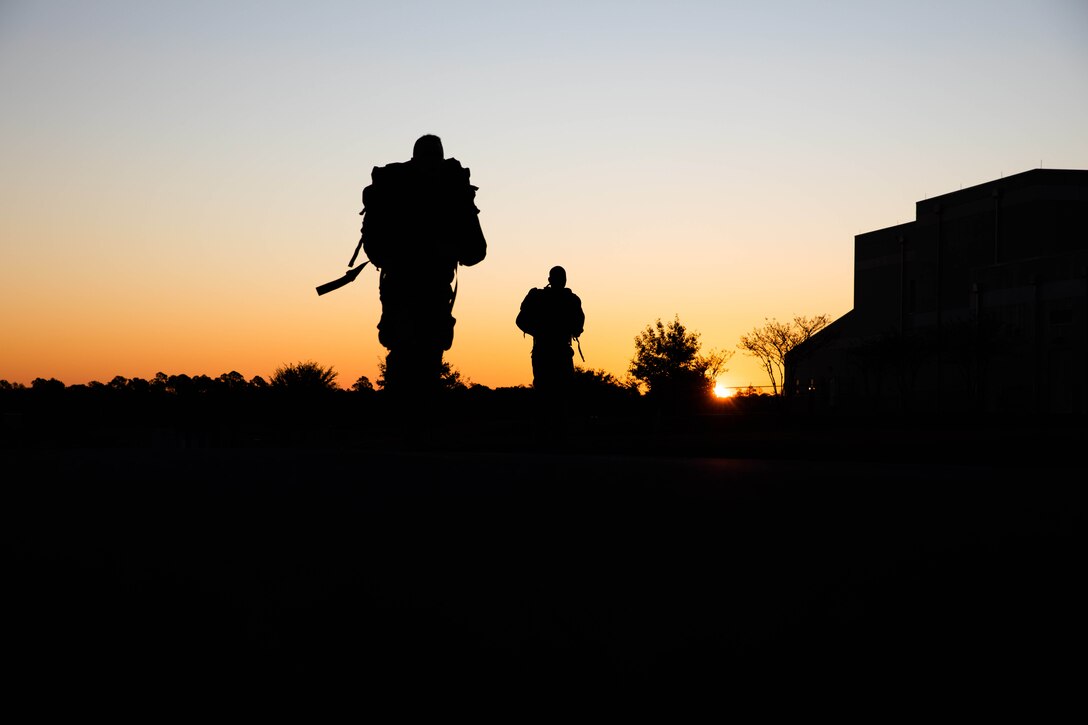 Two soldiers, seen in silhouette against a gold and blue sky, walk with rucksacks.