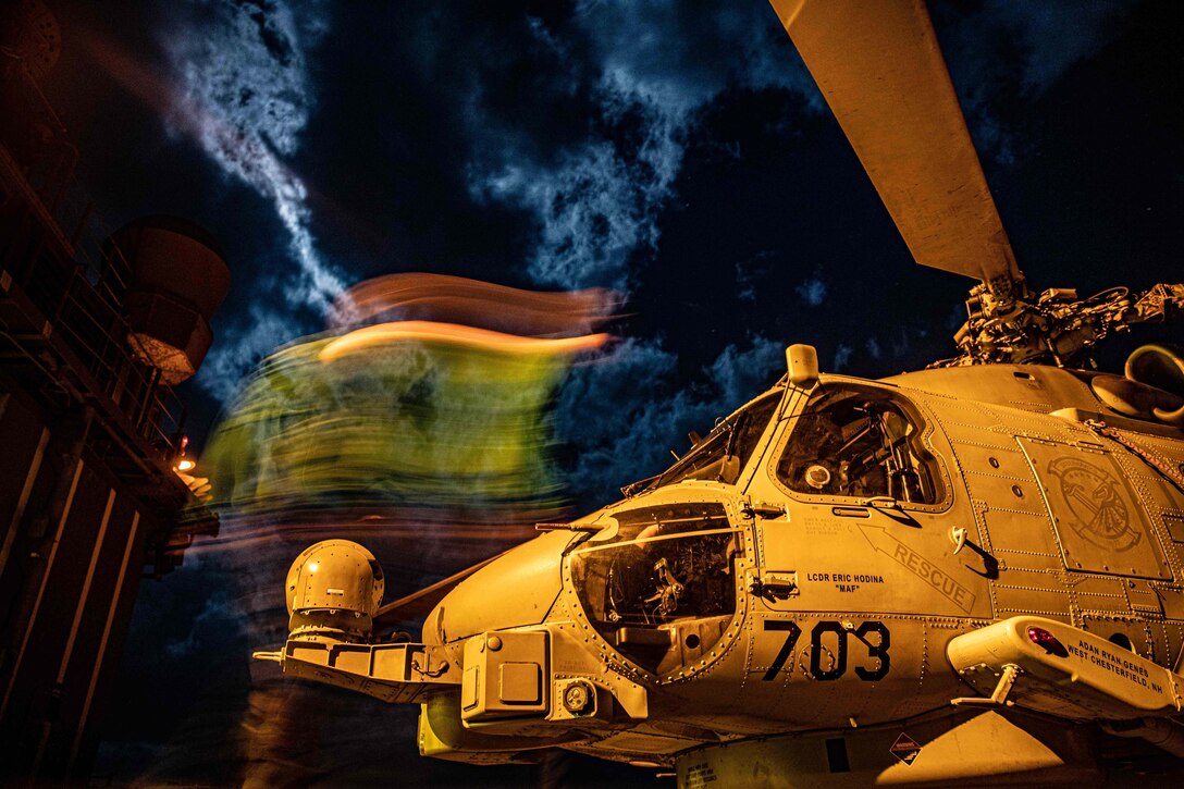 Red, yellow and green lights shift above a Seahawk aircraft on a ship's deck at night.