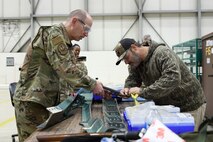 Photo shows two individuals tinkering with an aircraft part on a table.