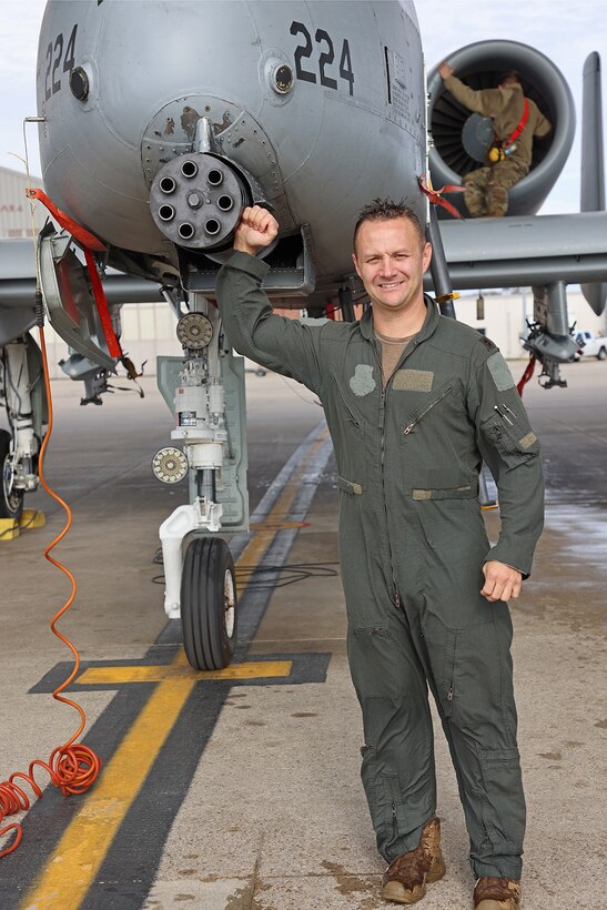 Pilot in jumpsuit stands next to A-10, hand on gun.