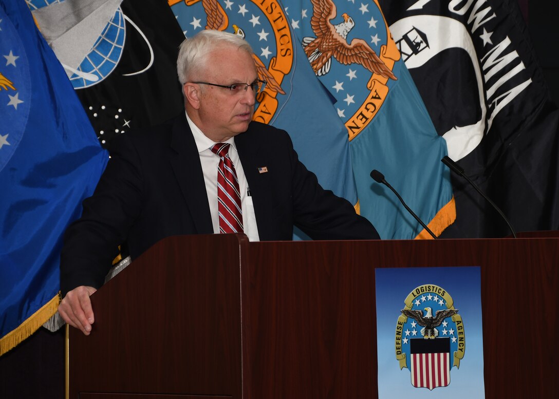Photo of a man at a podium. he has white hair and is in a dark suite.