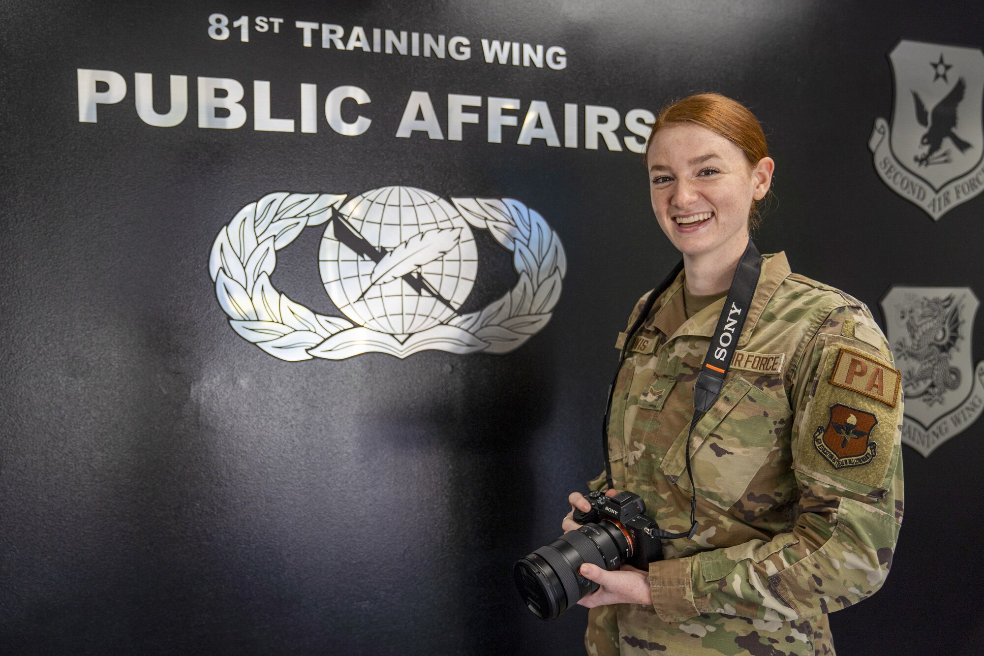 An Airman stands in front of a wall smiling and holding a camera.