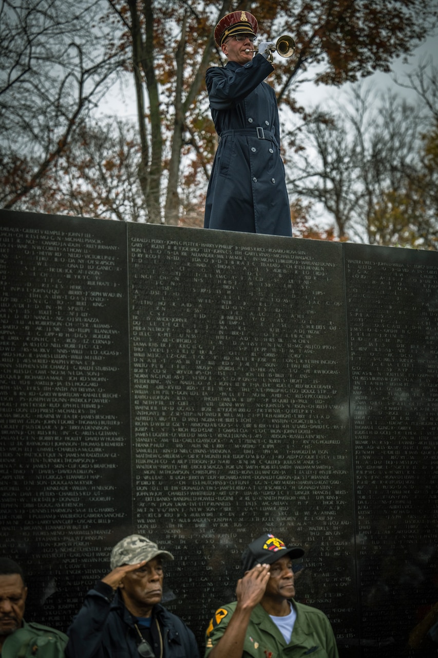 A man in uniform stands atop a wall and plays a bugle. Other men stand at the foot of the wall and salute.