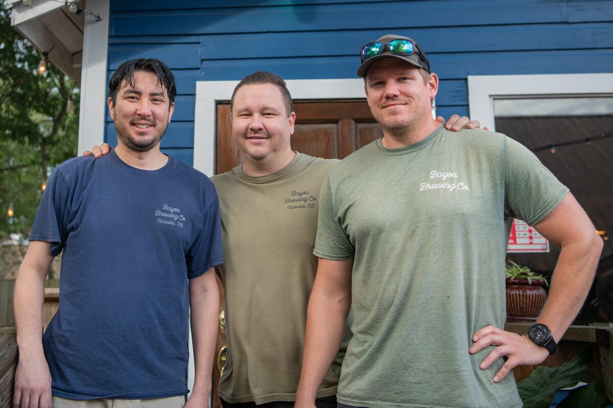 Three men pose for a photo in front of a storefront with hands on each others shoulders.