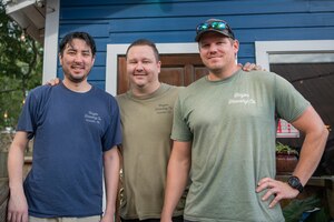 Three men pose for a photo in front of a storefront with hands on each others shoulders.