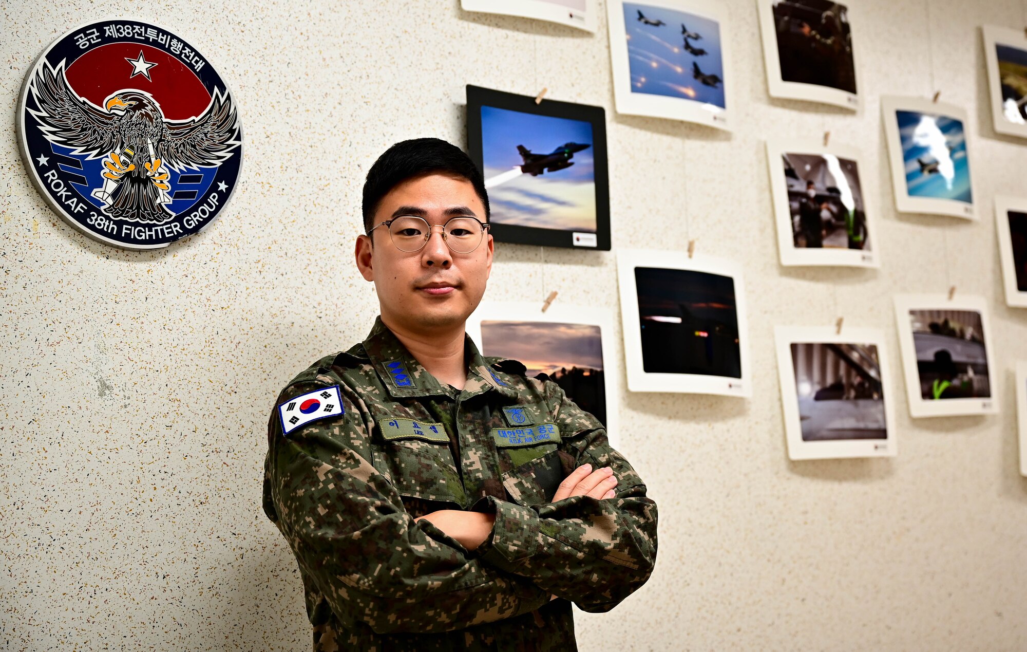 A ROK Airman stands in front of a photo wall with displayed images taken by ROKAF PA and a unit insignia on the wall