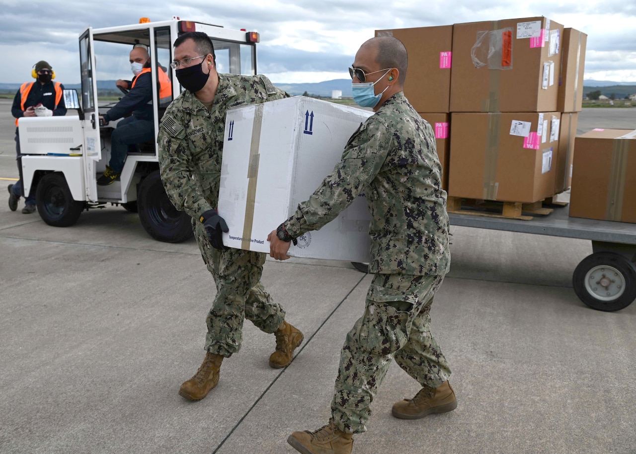 Two service member wearing COVID-19 masks carry a box while two others look on.