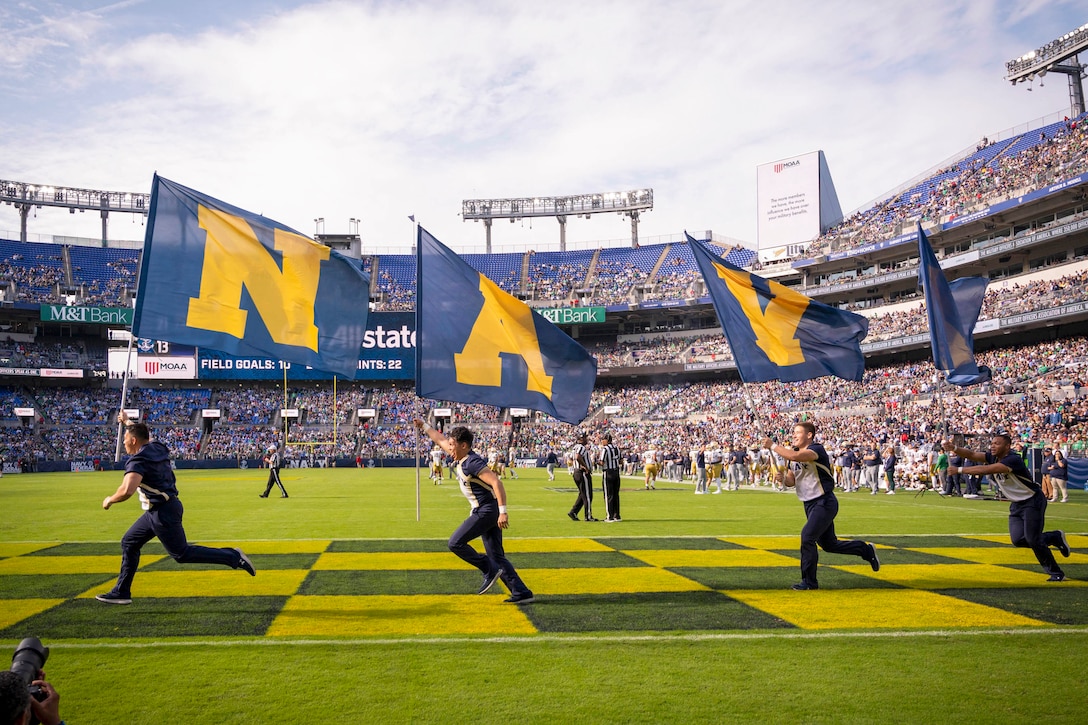 Cheerleaders holding flags run across a field during a football game.