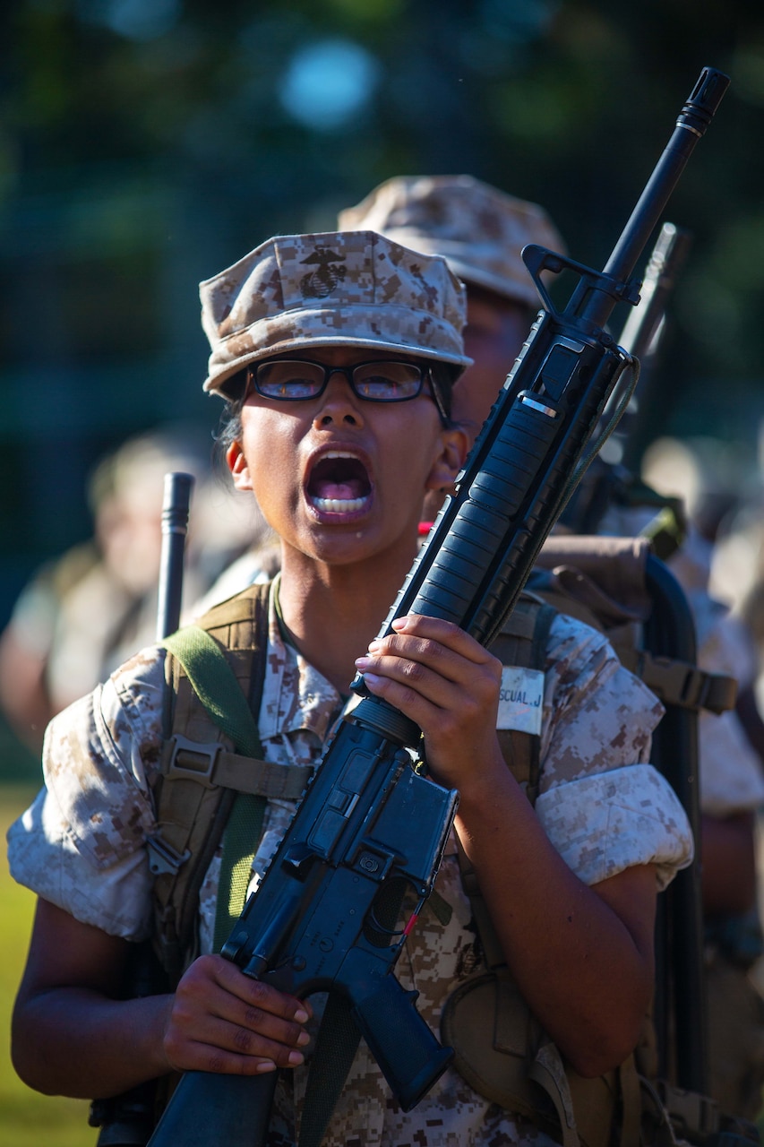 A Marine Corps recruit leading a line of others shouts a call while holding a training rifle.