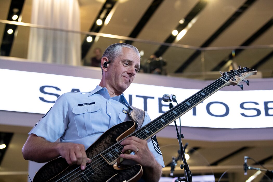 An Airman plays the bass guitar on a stage inside a sports arena.