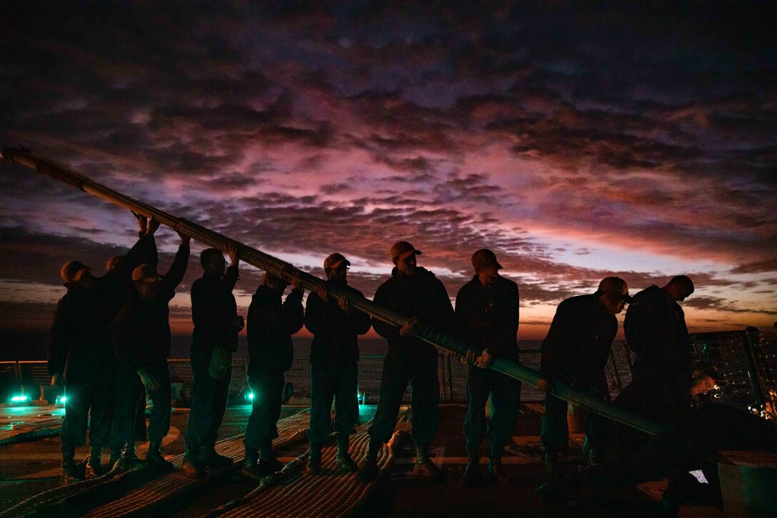 Sailors hoist a flag staff at night aboard a ship.