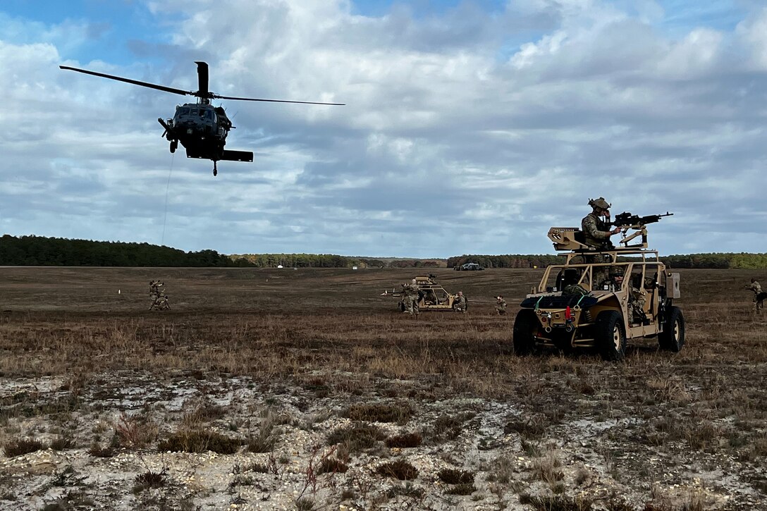 A helicopter hovers above the ground as troops  and military vehicles spread out below.