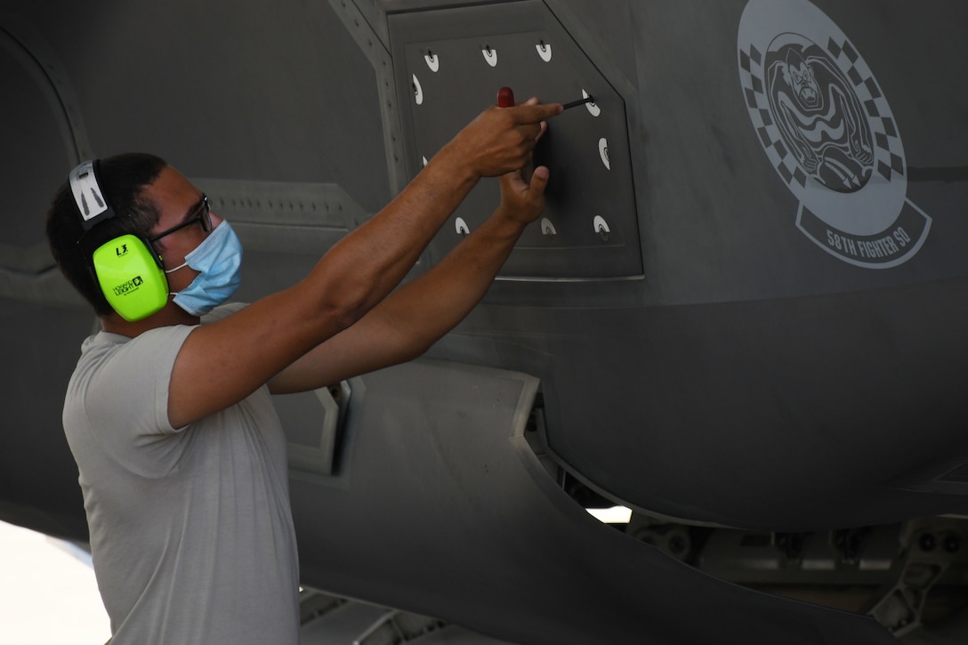Black male in a t-shirt uses a tool to close a panel on the outside of an aircraft