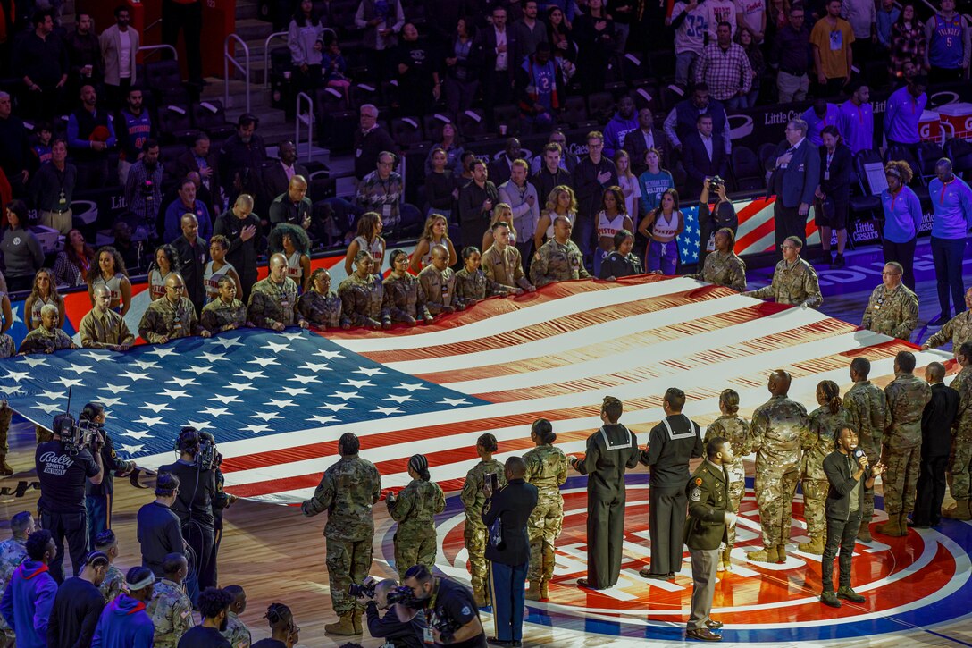 Service members hold an American flag on a basketball court.