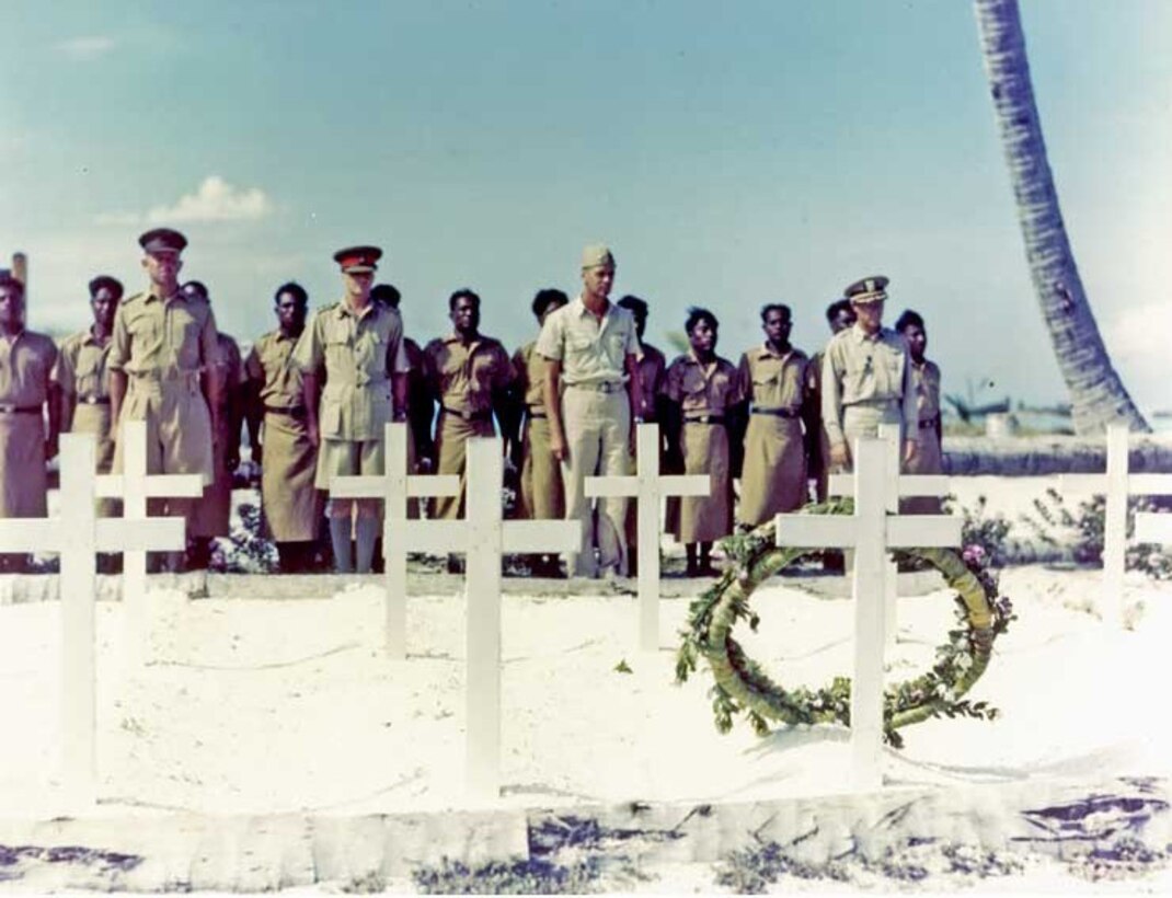 Several men stand in front of grave markers on a beach.