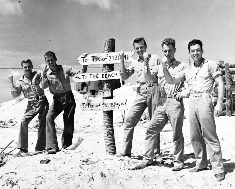 Five men hold their thumbs up by a signpost.
