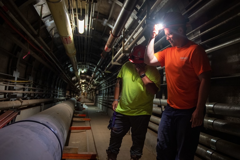 Two mean wearing hardhats stand near pipes that run down a long tunnel.