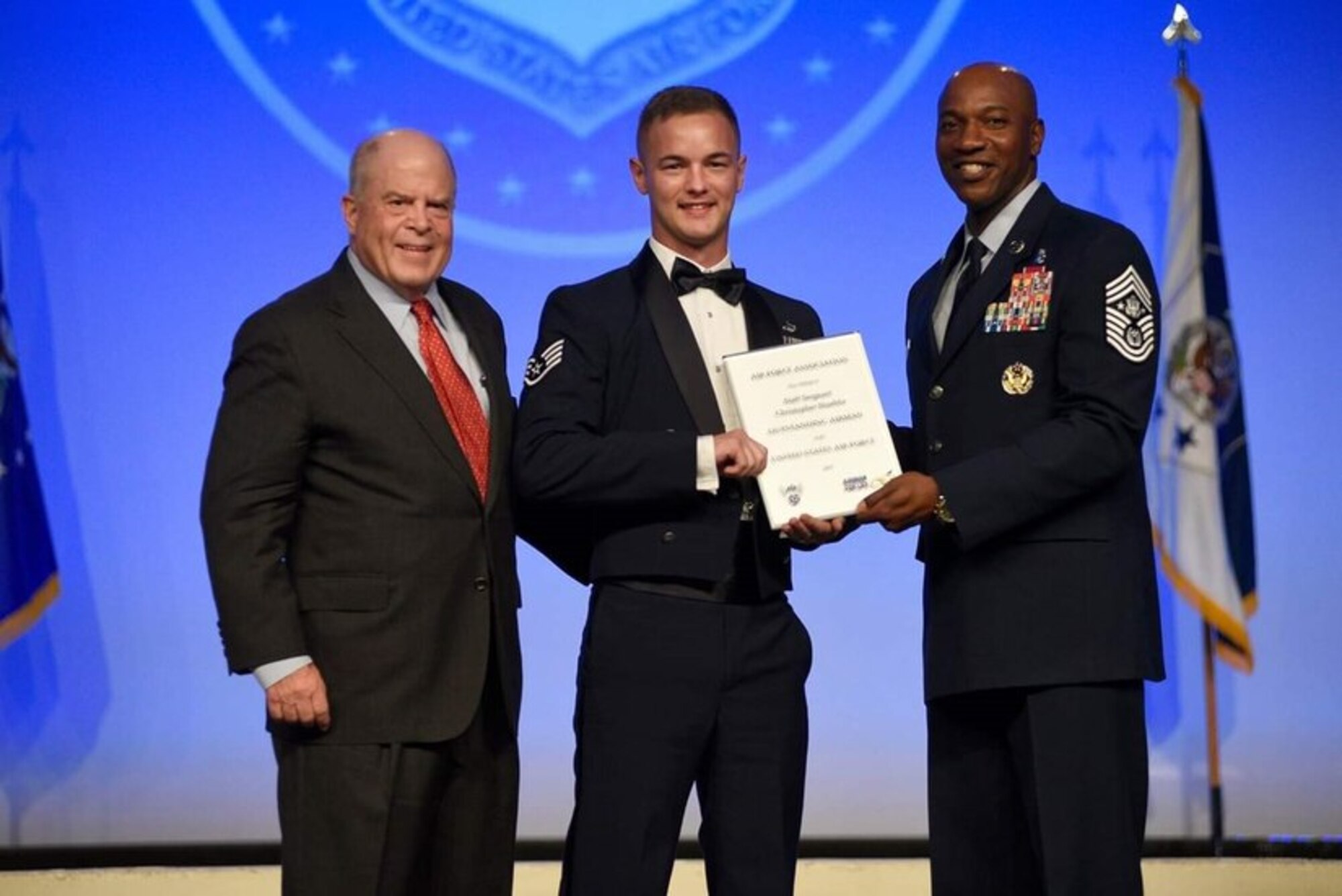 An Air Force Staff Sergeant poses for a photo with two other men. He holds a certificate and smiles.