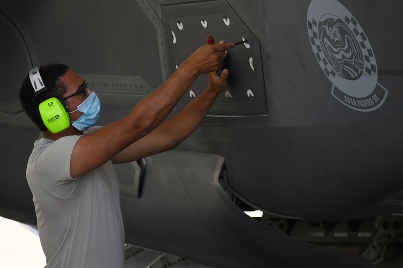 A man wearing a COVID-19 mask and hearing protection closes a panel on the side of an aircraft.