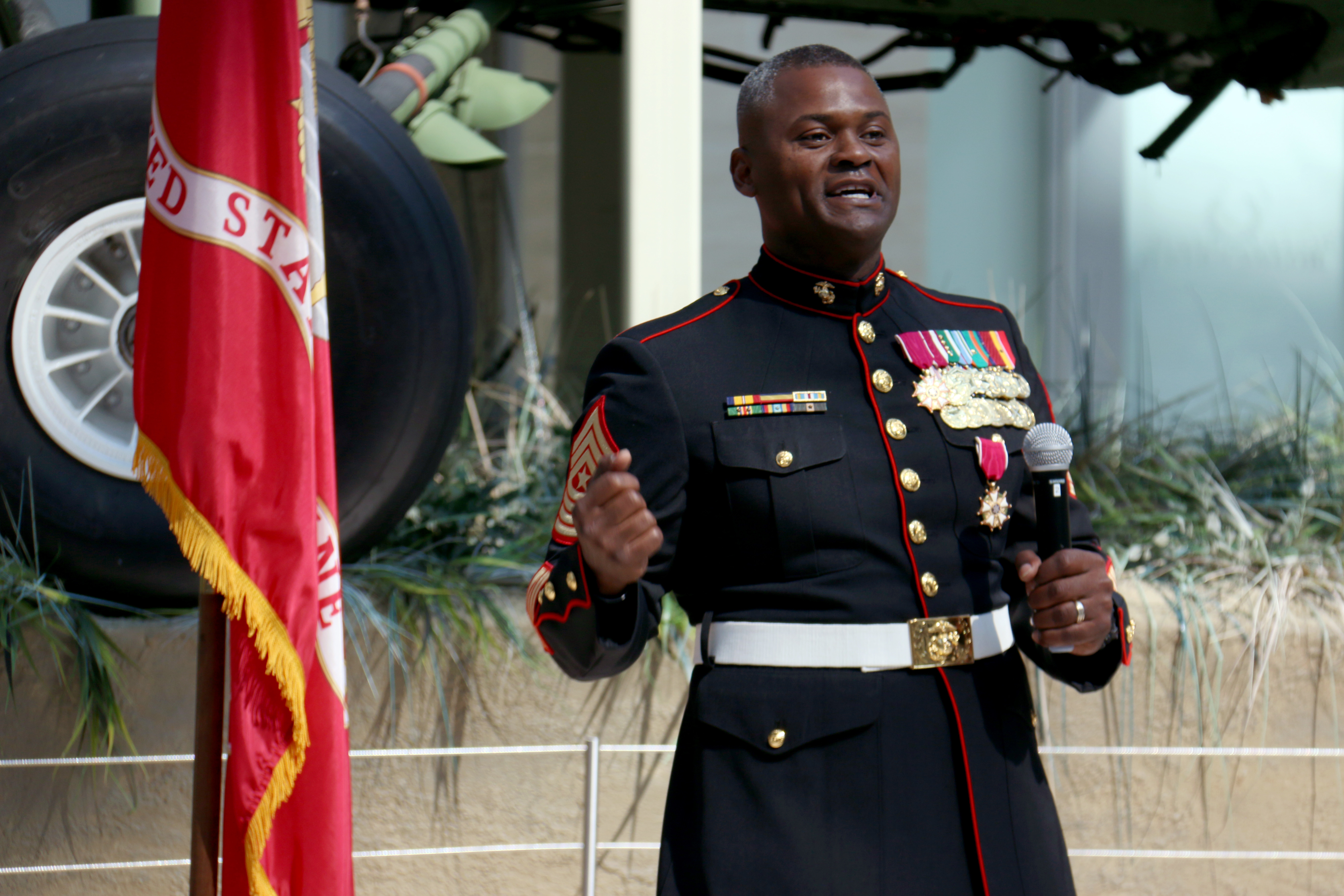 A Marine speaking with a microphone while standing in front of the Marine Corps flag.