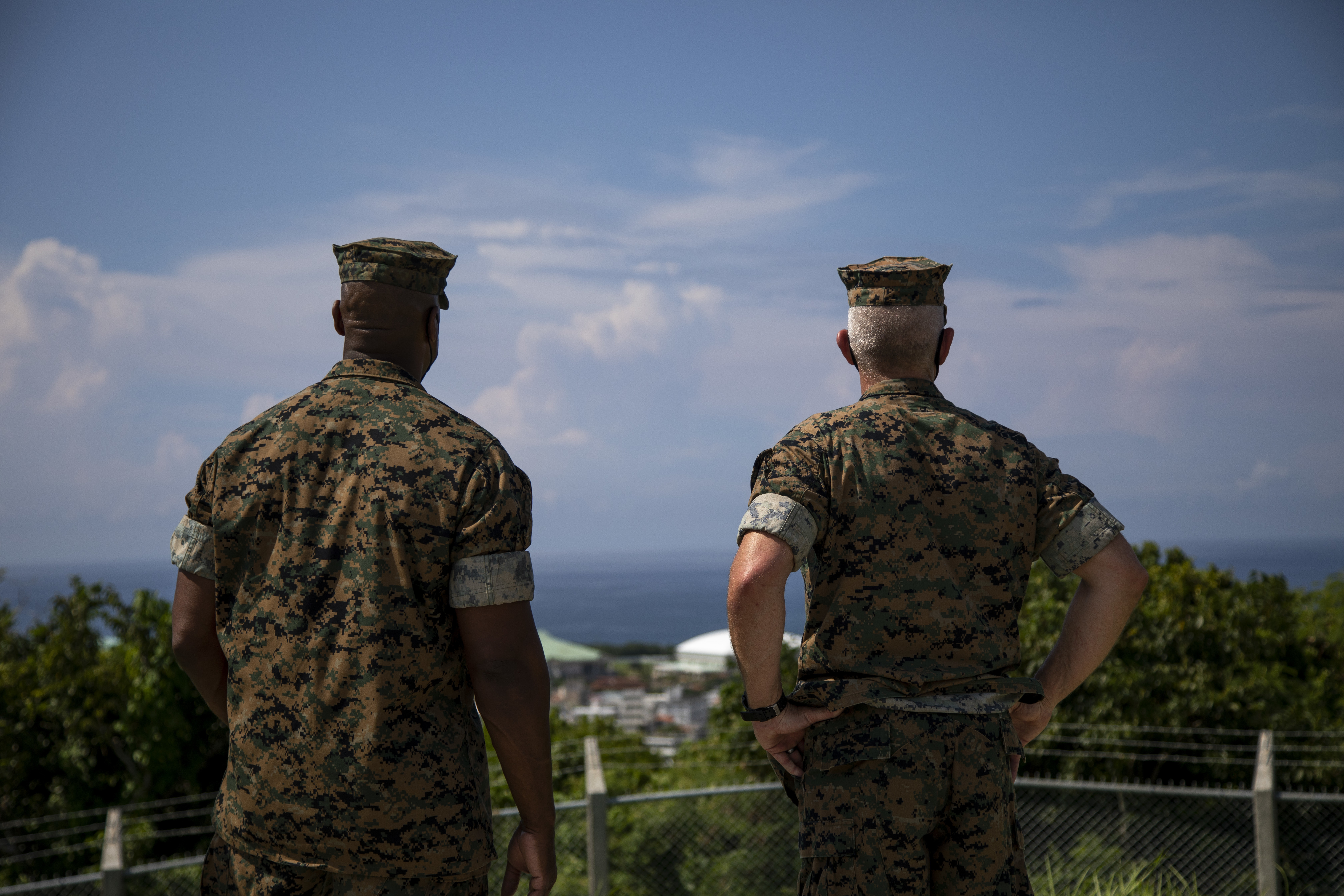 Two Marines in uniform with their backs to the camera, looking out at a clear blue sky.
