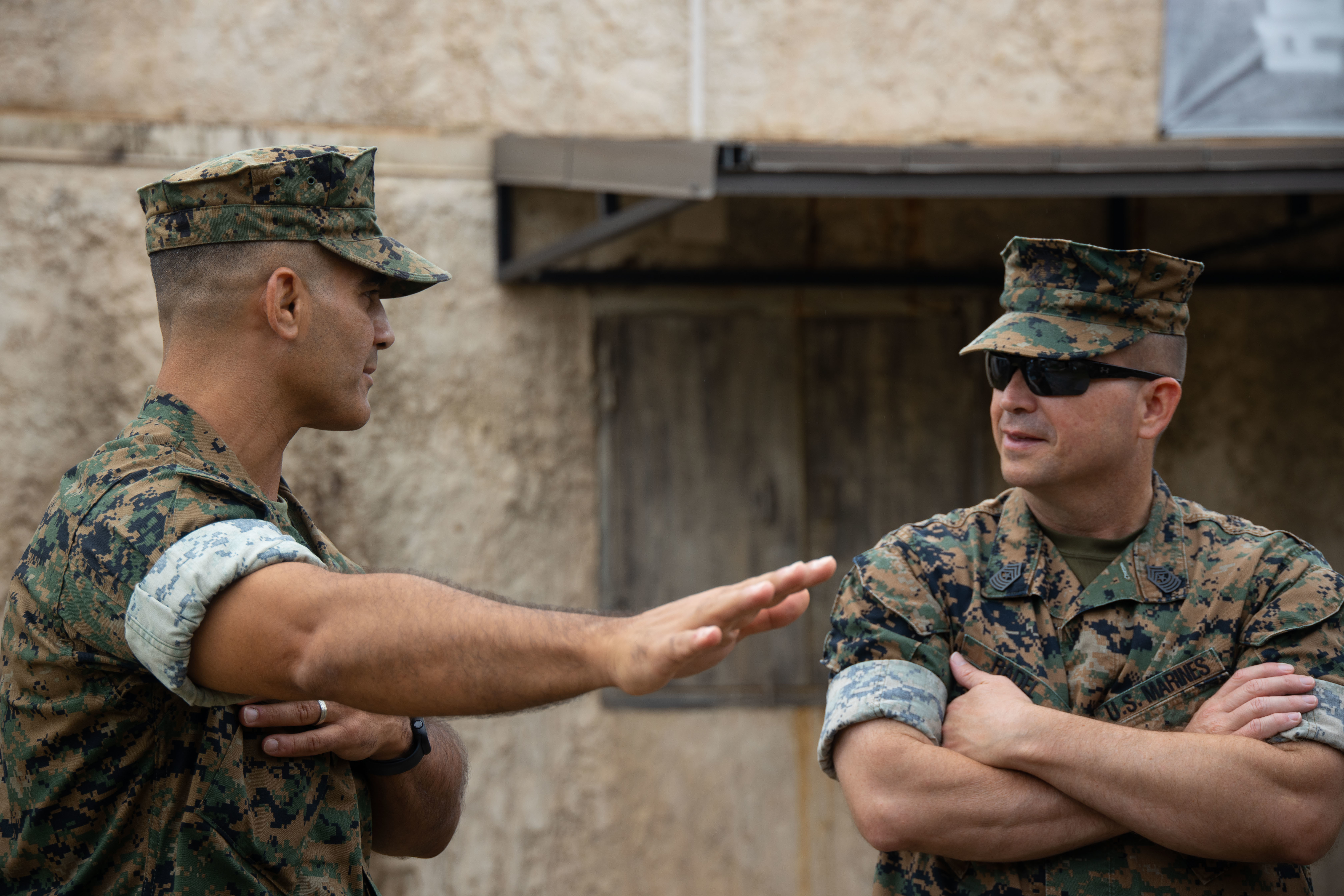 Two Marines talking outside a building, one with his arms crossed and the other pointing away.