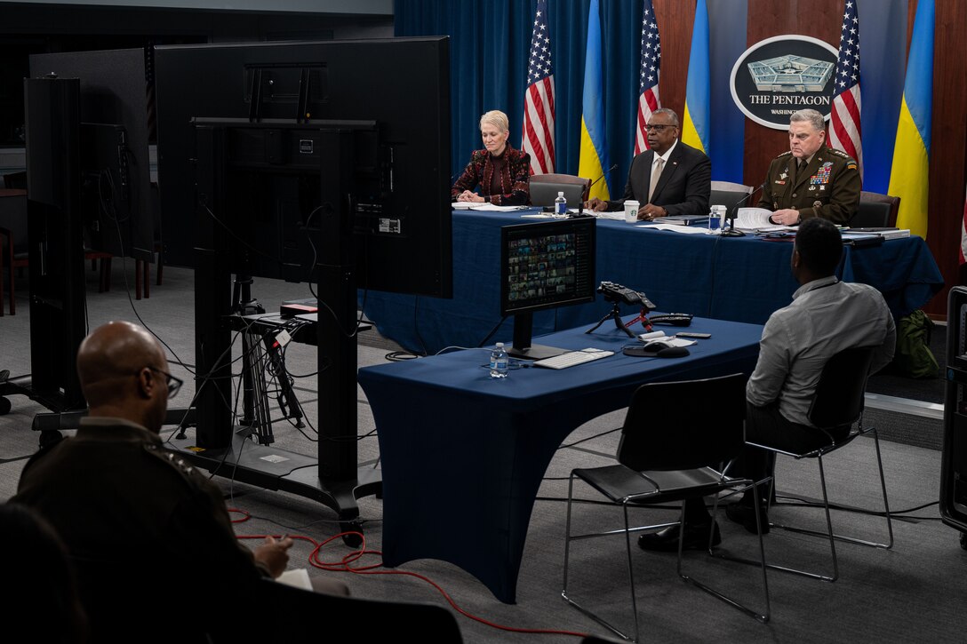 Two men and a woman sit at a table and look at a screen in front of them. Flags are in the background.