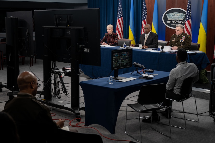 Two men and a woman sit at a table and look at a screen in front of them. Flags are in the background.