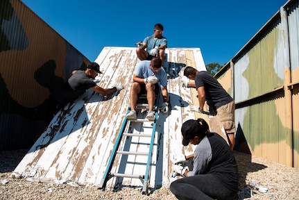 Five people are removing old paint from a large piece of wood