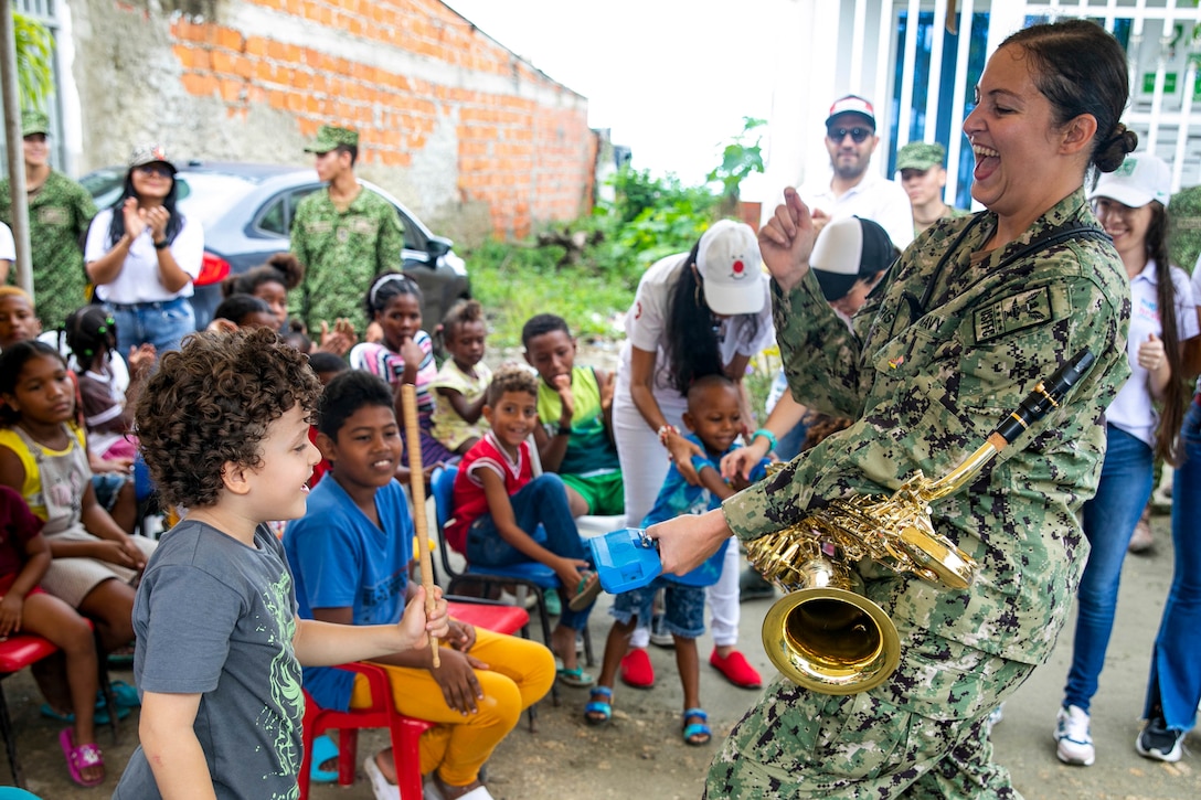 A laughing sailor wearing a saxophone holds out a block that a child hits with a stick as others watch.