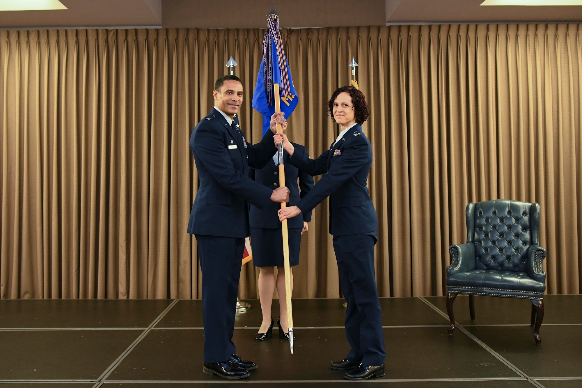 Two people in blue uniforms hold a guidon flag on stage during a ceremony.