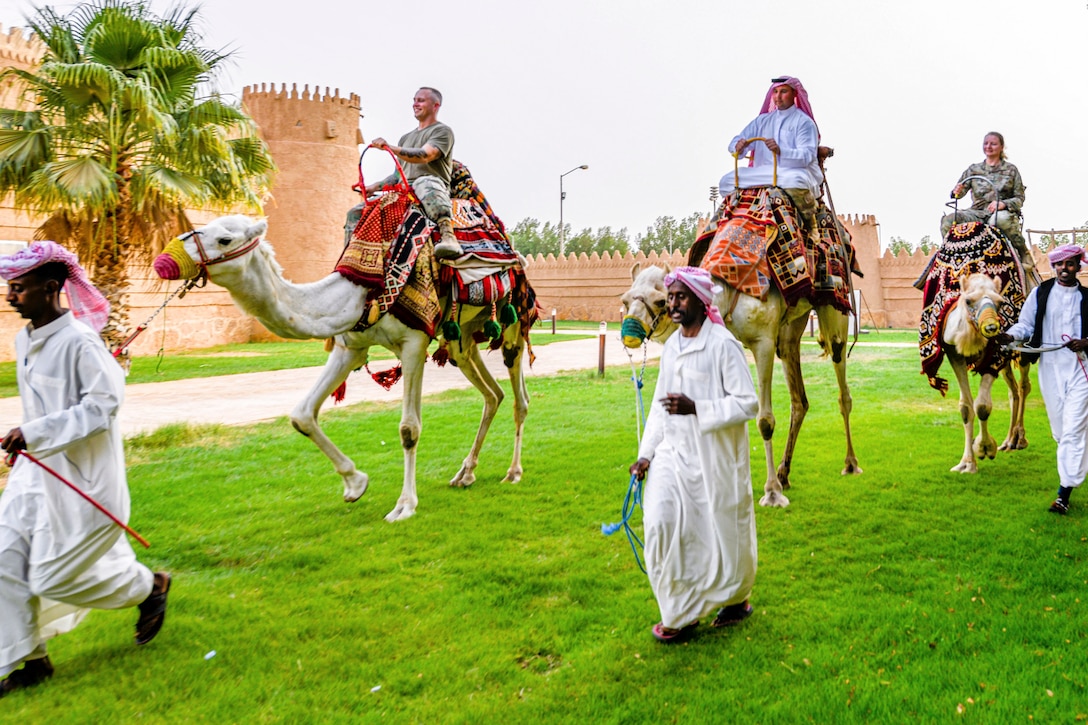 Service members ride camels by guides on a lawn.