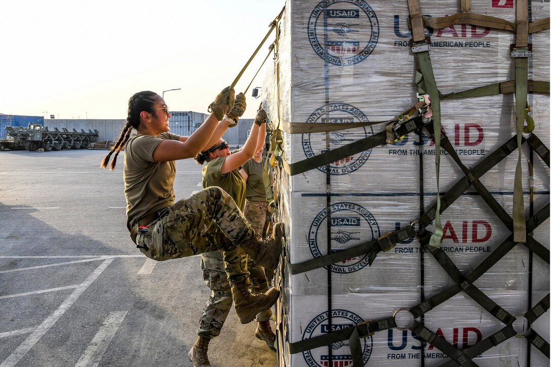 An airman pulls down on straps holding boxes on a pallet that sits on a flightline.