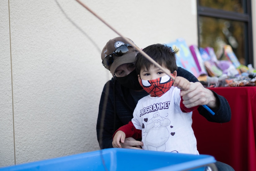 A small child holds a pole in the air and wears a Spiderman protective mask. An adult crouches behind the child and helps.