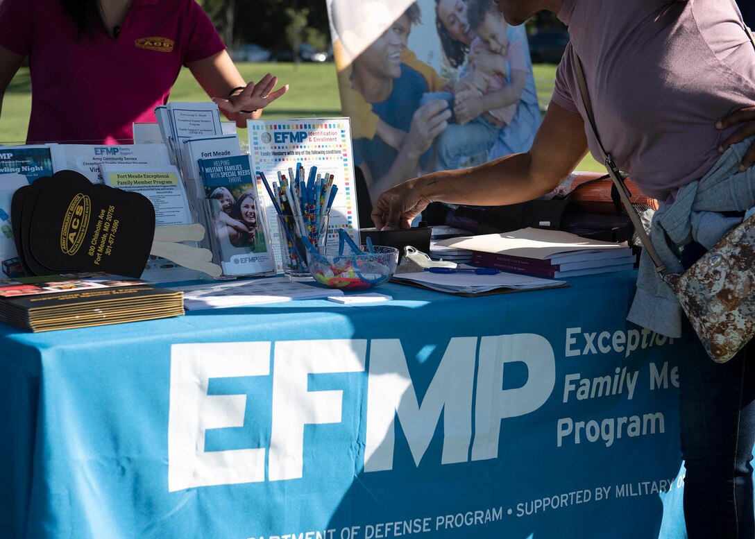Outdoors, a tablecloth has the letter EFMP on it. A person standing near the table selects some information from the table.