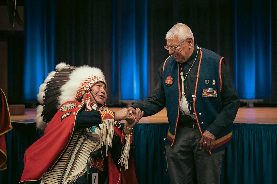 A retired Marine and a man dressed in traditional Native American clothing shake hands in an auditorium.