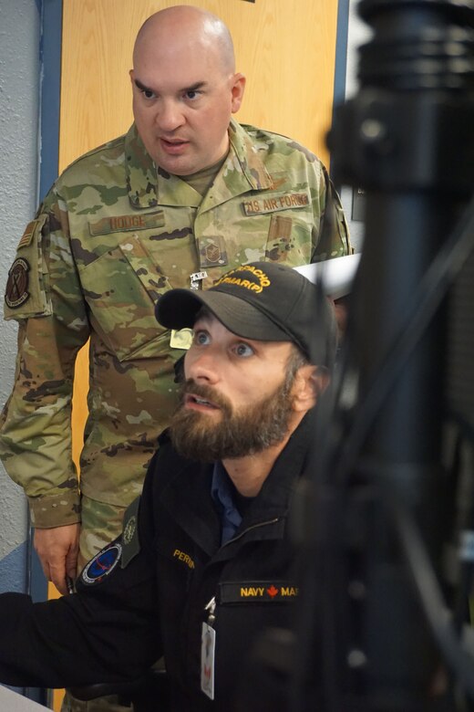 military member working at a computer, another military member is standing behind watching
