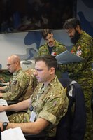 two military members sit working on computers while two additional military members stand behind them looking at a binder