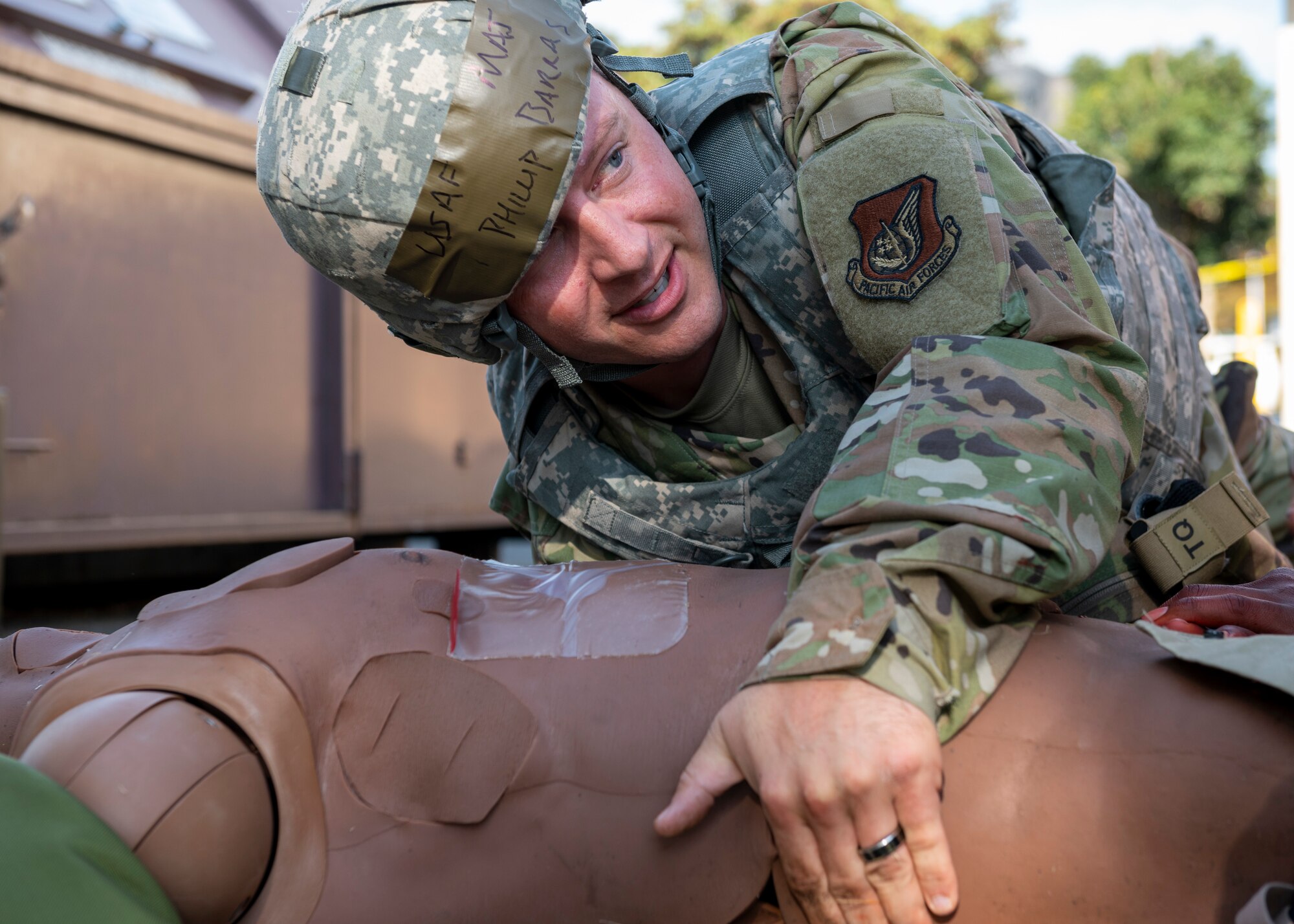 A man in combat gear attempts to roll over a medical training dummy