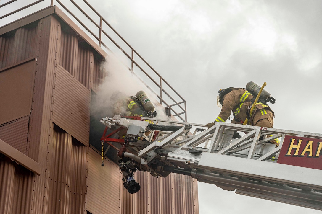 Two airmen dressed in firefighting gear climb a ladder into a building filled with smoke.