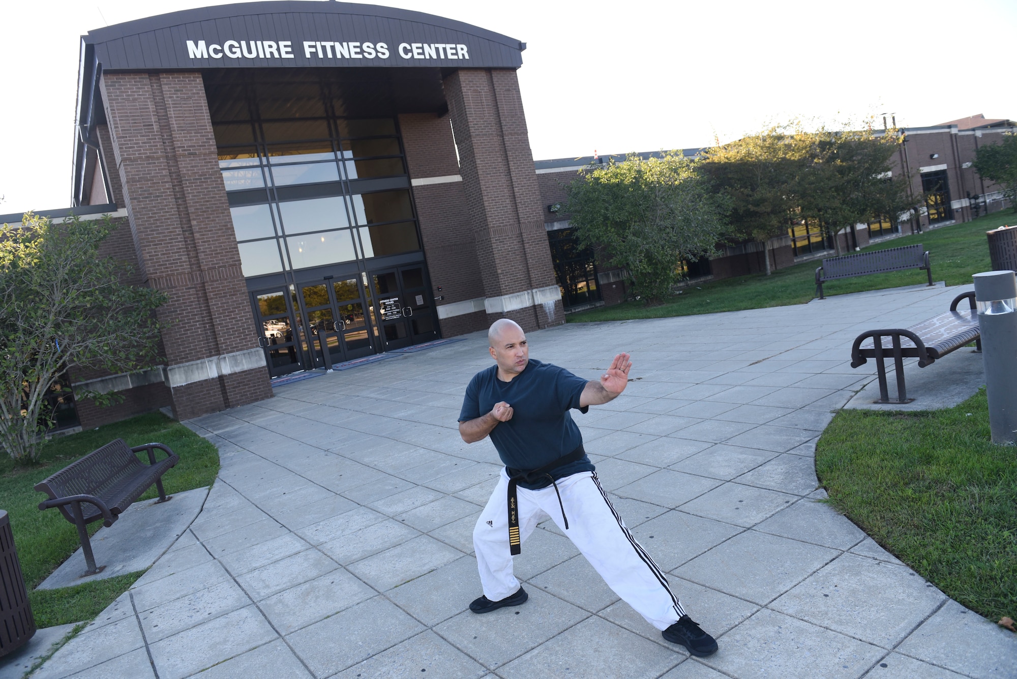 Staff Sergeant David Correa practices martial arts in front of the McGuire Fitness Center while wearing a black t-shirt, black belt, white pants, and black sneakers.