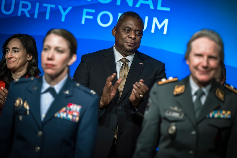 A man dressed in a business suit applauds as he stands behind women in uniform.